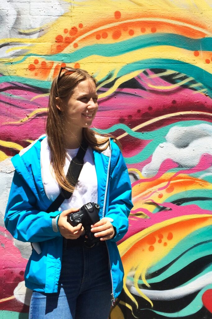 Junge Frau in Blauer Jacke vor einer bunten Graffity-Wand mit Kamera in der Hand