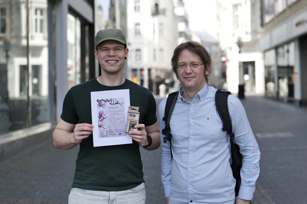 Jan Meyer und der Gewinner der Wildcard-Verlosung. XY hält die Urkunde und die EinwegKamera in das Bild.